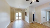 This single-floor residence offers an open and airy layout bathed in natural light. The main living area features warm hardwood flooring that extends throughout, contributing to a clean and cohesive aesthetic. Elevated ceilings enhance the sense of spaciousness, while a ceiling fan with a light fixture ensures year-round comfort. A decorative pendant chandelier adds a touch of elegance to the dining area.

A set of French doors serves as the focal point of the living space, opening to an outdoor balcony and filling the interior with abundant natural light. Transom windows above the doors further amplify the brightness of the room.

The kitchen is accessible via a convenient pass-through window with a breakfast bar, allowing for an open connection between the kitchen and living areas. Dark cabinetry in the kitchen provides a striking contrast to the neutral, warm-toned walls throughout the residence.

The overall color palette is soft and neutral, with beige walls creating a versatile backdrop suitable for a variety of interior styles. Floor vents are integrated discreetly into the layout. The space strikes a balance between functional design and inviting ambiance, making it a comfortable and well-appointed single-floor living environment.