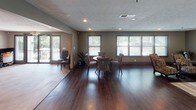 This single-floor community space is thoughtfully designed to blend comfort and functionality in an open, welcoming layout. Rich, dark hardwood flooring flows seamlessly throughout, anchoring the warm, neutral color palette of taupe and gray tones that define the space's sophisticated ambiance.

The main lounge area serves as the centerpiece, featuring a tile-surround fireplace with a sleek dark mantel adorned with decorative accents. Stylish seating, including bold animal-print accent chairs and a plush sofa, is arranged around a glass-top coffee table atop a neutral area rug, creating an inviting gathering space. Decorative framed artwork and large mirrors enhance the walls, adding depth and visual interest.

Adjacent to the lounge, a dedicated seating nook with a round table and chairs offers a quieter setting for smaller gatherings or individual use. Multiple windows with blinds line the walls, flooding the space with ample natural light and offering views of the surrounding greenery.

The kitchenette area is well-appointed with dark cabinetry, a crisp white countertop, pendant lighting, and a breakfast bar with barstool seating — ideal for casual refreshments. A coffee station and under-counter refrigerator add practical convenience. The textured ceiling with recessed lighting completes this polished, multifunctional space.