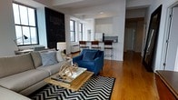 This single-floor space showcases an open-concept layout that seamlessly connects the living area and kitchen, creating a spacious and functional environment. The living area is anchored by warm hardwood floors and features a bold chalkboard accent wall that adds an urban, industrial character to the space. Large windows allow natural light to filter in, brightening the interior throughout the day.

The living area is generously proportioned, offering ample room for comfortable seating arrangements. A chevron-patterned area rug defines the lounge zone, complemented by a mix of contemporary and classic furnishings, including a plush gray sofa and a striking navy blue accent chair.

The kitchen is efficiently designed with clean white cabinetry and a peninsula counter with bar-height seating, providing casual dining options and additional workspace. Stainless steel appliances complete the modern aesthetic.

Structural beams and columns are subtly integrated into the design, hinting at the building's architectural heritage while maintaining a clean, contemporary feel. Overall, the space strikes a balance between modern styling and warm, inviting character.