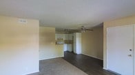 This single-floor space features an open-concept layout that seamlessly connects the living area to the kitchen. The living space is divided by flooring materials, with dark hardwood floors transitioning to carpeted areas, adding visual variety and defining different zones within the room. A ceiling fan with a light fixture is centrally positioned, providing both air circulation and overhead lighting.

The kitchen is situated at the rear of the open layout and features white cabinetry with under-cabinet range ventilation, white appliances including a range and refrigerator, and a clean, functional design. The cabinetry extends to the upper walls, maximizing storage. A textured popcorn ceiling runs throughout the space, and the walls are finished in a warm neutral tone that contributes to a light, understated ambiance. White baseboards provide a clean border throughout. The open flow between the living and kitchen areas makes the space feel connected and practical, suited for everyday living.