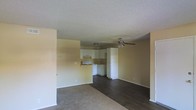 This single-floor space features an open-concept layout that seamlessly connects the living area to the kitchen. The living space is divided by flooring materials, with dark hardwood floors transitioning to carpeted areas, adding visual variety and defining different zones within the room. A ceiling fan with a light fixture is centrally positioned, providing both air circulation and overhead lighting.

The kitchen is situated at the rear of the open layout and features white cabinetry with under-cabinet range ventilation, white appliances including a range and refrigerator, and a clean, functional design. The cabinetry extends to the upper walls, maximizing storage. A textured popcorn ceiling runs throughout the space, and the walls are finished in a warm neutral tone that contributes to a light, understated ambiance. White baseboards provide a clean border throughout. The open flow between the living and kitchen areas makes the space feel connected and practical, suited for everyday living.