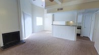 This two-story residence showcases a dramatic vaulted ceiling in the main living area, creating an expansive sense of height and openness. A ceiling fan is mounted at the peak, complementing the natural light that filters in through a large sliding glass door fitted with vertical blinds along one wall. The living area features neutral carpeting that extends throughout the main level, contributing to a clean, cohesive aesthetic.

A half-wall partition with white paneling defines the kitchen area, which includes a raised bar-height counter with a dark laminate surface — ideal for casual dining or meal prep. Under-cabinet lighting provides functional illumination in the kitchen space. Adjacent to the kitchen, a separate dining area is visible, highlighted by a chandelier that adds a decorative touch. The white-paneled walls throughout lend a bright, consistent finish to the interior.

The second floor is accessible via the interior layout, with a loft-style opening visible above, overlooking the main living space. The overall ambiance is airy and light-filled, with a simple, neutral palette that provides a versatile canvas throughout.