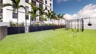 This outdoor gym in Miami, FL offers a well-designed open-air fitness environment that seamlessly blends functionality with an inviting urban atmosphere. The ground is covered entirely in vibrant artificial turf, providing a clean, low-maintenance surface ideal for a wide range of workout activities. At the center of the space stands a robust multi-functional fitness rig, equipped with suspension trainers, battle ropes, a punching bag, and pull-up bars, catering to strength, cardio, and functional training needs. The area is enclosed by sturdy stucco walls and sleek dark metal fencing, creating a secure and defined workout zone with a gated entry point for controlled access. Tall palm trees line one side of the perimeter, adding a tropical aesthetic and partial shade that is characteristic of Miami's outdoor spaces. The open-sky design ensures abundant natural light throughout the day, while sweeping views of Miami's dynamic city skyline serve as an inspiring backdrop. The surrounding urban landscape, visible beyond the perimeter walls, reinforces the gym's prime location in the heart of the city. This outdoor fitness space provides a comprehensive and energizing environment for users seeking an effective workout experience in the open air.