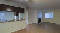 This single-floor space presents an open-concept layout that seamlessly connects the kitchen and living areas. The kitchen is defined by a pass-through counter with granite-style countertops, dark cabinetry overhead, and a pendant light fixture hanging above, providing both practical workspace and a visual boundary between the two areas. A mirrored backsplash adds depth to the kitchen space.

The living area is spacious and open, featuring wood-style laminate flooring that runs throughout, lending warmth and continuity to the space. A fireplace with a sleek black surround serves as a natural focal point along the wall, adding both a functional and aesthetic element to the room. Neutral-toned walls and white baseboards keep the interior feeling bright and clean.

A window fitted with vertical blinds allows natural light to filter into the living area, while a standard interior door leads to an additional room or utility space. The ceiling fan contributes to year-round comfort. Overall, the space maintains a straightforward, functional design with practical finishes well-suited for everyday living.