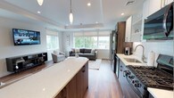 Unit 202 is a well-appointed 1-bedroom, 1-bathroom residence thoughtfully designed with a modern aesthetic and functional open-concept layout. The kitchen serves as a central hub of the living space, anchored by a large island with a sleek white quartz countertop and rich wood cabinetry. Stainless steel appliances, including a gas range and refrigerator, complement the two-tone cabinetry featuring gray lower cabinets and warm wood upper accents. Recessed lighting and a stylish pendant light above the island add both ambiance and practicality. The kitchen flows seamlessly into the living area, which is bathed in natural light from a generous bank of windows offering views of the surrounding greenery. A tray ceiling adds architectural interest and a sense of height to the living space. Warm hardwood-style flooring runs throughout, grounding the open layout with a cohesive, inviting feel. The overall design balances clean, contemporary lines with warm tones, creating a comfortable and stylish living environment. This single-floor unit offers a well-organized and efficiently designed space ideal for modern living.
