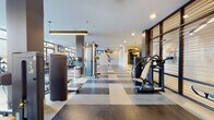 This contemporary fitness center is thoughtfully designed to accommodate a wide range of workout styles across a single, expansive floor. The space is defined by its striking checkerboard-patterned tile flooring in contrasting shades of dark charcoal and light grey, lending the facility a clean, polished aesthetic throughout.

The cardio zone lines the perimeter near large floor-to-ceiling windows, which flood the space with natural light and offer open views of the surrounding outdoor area. This section is outfitted with rowing machines, stationary bikes, ellipticals, and treadmills. Multiple wall-mounted televisions are positioned throughout for entertainment during workouts.

The strength training area features a comprehensive selection of equipment, including cable machines, multi-functional racks, free weight benches, and a dumbbell rack. Full-length mirrors line the walls of this section, enhancing the sense of space and allowing users to monitor their form. A dedicated functional training zone includes plyometric boxes, a punching bag station, and suspension training equipment.

The facility also includes a glass-enclosed entry vestibule with sleek black steel framing, a wall-mounted cubby storage unit, and an accessible dual water fountain station set against a textured tile accent wall. Ceiling fans, recessed lighting, and linear pendant fixtures ensure consistent, comfortable illumination throughout.