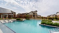 This outstanding outdoor amenity space centers around a generously sized resort-style swimming pool with crystal-clear blue water, designed to evoke a relaxed, vacation-inspired atmosphere. The pool deck is finished in large-format stone tiles and is lined with rows of chaise lounges accompanied by red-accented umbrellas, offering ample space for sunbathing and outdoor relaxation. Integrated planting beds featuring ornamental grasses rise directly from the pool's edge, adding a natural, landscaped aesthetic that softens the hardscape surroundings.

A standout architectural feature is the clubhouse building, which showcases a striking board-and-batten white facade with a steeply pitched black metal roof and expansive floor-to-ceiling windows, contributing a modern farmhouse aesthetic to the overall design. Adjacent to the pool, a thoughtfully designed wood pergola with horizontal slatted screens provides semi-private shaded retreats, each outfitted with individual lounge chairs for a more intimate outdoor experience. The tiled flooring extends seamlessly throughout these shaded nooks, maintaining a cohesive and polished look.

The surrounding multi-story residential buildings feature a mix of neutral stucco, warm-toned brick, and dark trim accents, with private balconies overlooking the pool area. The entire amenity space is enclosed by sleek black metal fencing, bordered by manicured green lawns and young trees that enhance the open, airy ambiance of this well-appointed outdoor retreat.