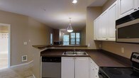 This single-floor residence offers an open-concept layout that connects the kitchen and living area in a cohesive, functional design. The kitchen is well-appointed with crisp white cabinetry providing ample storage, dark countertops that create a striking contrast, and a double stainless steel sink positioned along the counter. A stainless steel dishwasher and additional appliances complement the space. Recessed lighting and a stylish pendant light fixture hanging above the kitchen area provide layered illumination throughout. The kitchen features a wrap-around counter that serves as a natural divider between the cooking area and the adjacent living space. The living area beyond is bathed in natural light from a set of windows that look out to a lush, tree-lined exterior, creating a pleasant connection with the outdoors. Warm, neutral-toned walls throughout give the space a cohesive and inviting atmosphere. A second pendant light fixture is visible in the living area, further enhancing the warm ambiance. A microwave is also integrated into the kitchen setup, adding to the functional appeal of this well-designed, single-level home.