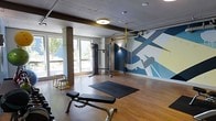This single-floor fitness center offers a thoughtfully designed workout space that balances functionality with an energizing aesthetic. The room is anchored by a striking large-scale graphic mural on the feature wall, rendered in bold shades of blue, white, and yellow, creating a dynamic and motivating atmosphere for exercise.

The space is outfitted with a variety of training equipment, including a multi-function cable machine, a TRX suspension training system, an adjustable workout bench, and a plyo box, providing options for strength, functional, and bodyweight training. A large exercise mat laid on the hardwood floor offers dedicated space for stretching, floor exercises, or core work.

Structural concrete columns add an industrial character to the interior, complemented by exposed ceiling elements and ductwork that lend a loft-like quality to the space. Recessed and flush-mount ceiling lighting ensures the room is well-lit throughout.

Natural light streams in through generously sized windows along one wall, offering views of surrounding trees and greenery, contributing to a bright and refreshing workout environment. The warm hardwood flooring ties the space together, creating a clean, inviting foundation for all fitness activities. Overall, this fitness center is designed to be both practical and visually engaging.