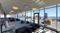 This sophisticated single-floor lounge space is defined by an expansive wall of floor-to-ceiling glass windows and doors that flood the interior with natural light and frame breathtaking panoramic views of the surrounding city skyline and a shimmering body of water in the distance. The open, airy layout is thoughtfully arranged with multiple distinct seating clusters, offering a variety of social settings within a single space. Deep, low-profile sofas and oversized bean bag-style poufs in dark tones provide a relaxed, contemporary seating option, while accent chairs in bold red add a vibrant contrast to the neutral palette. The flooring features a patterned area rug that grounds the central gathering zones. Warm circular ceiling fixtures cast a soft ambient glow, complementing the abundant daylight streaming through the glass facade. A door along the window wall opens directly onto an outdoor terrace, seamlessly extending the space and further enhancing its connection to the urban landscape beyond. The overall ambiance is modern and elevated, combining clean architectural lines with comfortable, design-forward furnishings to create a versatile space ideal for both relaxed gatherings and more structured social interaction.