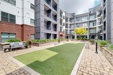 Located on Ponce de Leon Avenue in Atlanta's vibrant Midtown neighborhood, this inviting outdoor courtyard serves as a central amenity space framed by a multi-story residential building. The architecture blends classic red brick with contemporary gray paneling, creating a modern yet warm aesthetic that carries throughout the surrounding façades. Private balconies with black iron railings overlook the courtyard at each level, reinforcing the sense of an enclosed, community-centered space.

At ground level, the courtyard is thoughtfully designed with a combination of paver stone walkways and a well-maintained artificial turf lawn, offering both practical and recreational areas. Stone-capped raised planter beds line the perimeter and central areas, filled with ornamental grasses, flowering plants, and a standout mature tree that anchors the space with natural beauty. Decorative river rock accents complement the planters and add textural variety throughout the landscaping.

Outdoor seating areas with bistro-style tables and chairs are positioned throughout the courtyard, encouraging gathering and relaxation. Pergola structures provide shaded spots for residents to enjoy the outdoors. The overall ambiance is green, open, and serene—offering a tranquil retreat within an urban setting.
