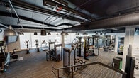 Located on Reseda Boulevard in Northridge, Los Angeles, this single-floor fitness center offers a generously sized exercise space designed to support a comprehensive workout experience. The facility's industrial-inspired aesthetic is defined by an exposed ceiling with visible ductwork, structural concrete columns, and dark pendant lighting that creates a focused, energizing atmosphere throughout.

The exercise room is thoughtfully equipped with a wide range of cardio machines, including treadmills, ellipticals, and rowing machines, all positioned along the perimeter to take advantage of large floor-to-ceiling windows that flood the space with natural light and offer views to the outside. The strength training area features a multi-station cable machine, free weight racks stocked with dumbbells and kettlebells, weight benches, and additional resistance equipment.

Durable, striped carpet flooring in earthy tones adds both visual interest and functional comfort underfoot. A central staircase with metal railings provides access between levels of the broader facility. A vibrant Northridge-themed mural anchors one wall, adding a sense of local identity and character. The open layout ensures ample circulation space between equipment zones, accommodating multiple users simultaneously with ease.
