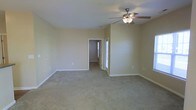 This single-story space presents a generously sized main living area finished with wall-to-wall carpeting in a neutral beige tone, contributing to a warm and understated ambiance throughout. The room is painted in soft, muted green-beige tones that complement the light-colored carpet and white baseboards, creating a cohesive and calming aesthetic. A ceiling fan with an integrated light fixture is centrally mounted, providing both air circulation and ambient lighting. The layout offers an open feel with clean sightlines across the room. Toward the rear, a doorway leads to an additional room, suggesting a connected layout that adds to the overall functionality of the space. A door with a glass panel, visible in the background, allows natural light to filter into the area, enhancing the brightness of the interior. The walls are smooth and feature minimal architectural detailing, maintaining a simple and practical design throughout. White baseboards run along the perimeter, providing a clean contrast to the carpeted floors. Overall, the space presents a straightforward, move-in-ready environment well-suited for comfortable everyday living.