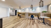 Located at 2840–2850 Carlow Rd in Victoria, BC, this single-floor space seamlessly blends a welcoming entrance lobby with a modern café-style lounge. The entrance area features large, black-framed windows that flood the space with natural light, large-format white tile flooring, and a built-in bar counter with seating along a wall adorned with a striking botanical mural and warm wood slat paneling. Glass-panel doors provide a clear view to the outdoors, reinforcing the bright and open atmosphere. Transitioning into the interior lounge, the flooring shifts to warm-toned wood-look planks that carry a casual, inviting feel throughout. A built-in Starbucks beverage station serves as a central anchor, featuring a sleek black and natural wood cabinet design with full coffee service equipment. Adjacent to the station, a comfortable seating area offers cushioned booth-style banquettes framed in natural wood, paired with bistro tables and contemporary black chairs. Decorative cage-style pendant lights hang above, and round mirrors mounted on the walls add depth and a polished finishing touch. The overall design balances clean, modern lines with warm natural materials, creating a functional yet stylish communal space well-suited for daily use.