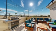 Located at 819-825 D Ave in National City, California, this shared outdoor amenity space is part of a modern multi-story residential building. The property features two distinct outdoor areas designed for communal gathering and relaxation. The rooftop terrace is the standout highlight, offering expansive panoramic views of the National City skyline, surrounding neighborhoods, and distant mountains under the open Southern California sky. This sun-drenched space is equipped with a stainless steel gas BBQ grill, teak wood dining and lounge furniture, a fire pit, and festive string lights that create an inviting atmosphere for both daytime and evening use. A covered ground-level courtyard complements the rooftop area, featuring clean white stucco walls, decorative snake plant planters in modern white rectangular pots, and a sheltered corridor connecting the building's units. The courtyard benefits from natural light filtered through the overhead structure, maintaining a bright and airy feel. Together, these outdoor spaces reflect a contemporary design aesthetic — clean lines, a neutral white palette, and thoughtfully curated furnishings — offering residents a functional and social outdoor environment in the heart of National City.