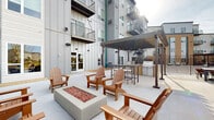 Located at 14211 Akron Ave in Rosemount, Minnesota, this thoughtfully designed outdoor amenity space serves as a central gathering hub for the surrounding residential community. The expansive concrete patio is anchored by a modern fire pit surrounded by classic wooden Adirondack chairs, creating an inviting lounge area ideal for relaxation and social gatherings.

A standout feature of the space is a sleek, dark-framed pergola that provides partial shade over a fully equipped outdoor kitchen. The kitchen station includes multiple gas grills, a prep counter with bar-height seating, and accent brick detailing — making it well-suited for outdoor dining and entertaining. String lights are draped across the pergola, adding ambiance for evening use.

The open patio extends along the building's exterior, offering clear sightlines to the surrounding neighborhood and tree-lined landscape beyond the perimeter railing. A secondary seating area near the building's edge provides a quieter retreat with views of the greenery and adjacent balconies above.

The overall design blends contemporary architecture — featuring mixed metal, panel siding, and large windows — with functional, resident-focused outdoor amenities. The space is fully open to the sky, maximizing natural light and creating an airy, welcoming atmosphere year-round.