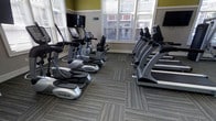 This fitness center offers a clean, purpose-built environment designed to support a comprehensive cardio workout routine. The space is outfitted with a lineup of True and Cybex branded cardio machines, including ellipticals and treadmills, arranged in organized rows to maximize the available floor area. Durable, patterned carpet tiles cover the floor throughout, providing both comfort underfoot and a polished aesthetic. Large windows fitted with adjustable blinds line the walls, allowing ample natural light to fill the room while offering views of the surrounding exterior. Wall-mounted flat-screen televisions are positioned at multiple angles, ensuring visibility from any machine and enhancing the workout experience. The walls feature a neutral two-tone color scheme with a warm green upper section and crisp white trim, giving the space a fresh and welcoming feel. The room's layout is thoughtfully organized, with sufficient spacing between machines to allow ease of movement and a comfortable workout environment. Overall, this fitness center presents a well-maintained, functional space equipped to meet the cardio fitness needs of its users.