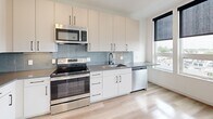 Located at 152 Albion St in Wakefield, Massachusetts, this single-floor unit presents a clean, contemporary design with consistent light wood flooring and crisp white walls throughout. The open-concept kitchen is a standout feature, equipped with stainless steel appliances including a French door refrigerator with ice maker, an electric range, an over-the-range microwave, and a dishwasher. White flat-panel cabinetry with sleek matte black hardware extends to the ceiling, providing ample storage, while a light gray subway tile backsplash and dark countertops lend a polished, modern aesthetic. A window alongside the kitchen brings in natural light and outdoor views. The living area is bright and open, featuring large windows fitted with roller shades that offer views of the surrounding neighborhood. The bathroom is thoughtfully designed with hexagonal tile flooring, a white undermount sink set in a light wood vanity with black fixtures, a large mirror, and a tiled shower. A dedicated closet provides additional storage. The overall ambiance is modern and minimalist, with neutral tones and quality finishes creating a cohesive and functional living environment.