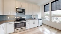 Located at 152 Albion St in Wakefield, Massachusetts, this single-floor unit presents a clean, contemporary design with consistent light wood flooring and crisp white walls throughout. The open-concept kitchen is a standout feature, equipped with stainless steel appliances including a French door refrigerator with ice maker, an electric range, an over-the-range microwave, and a dishwasher. White flat-panel cabinetry with sleek matte black hardware extends to the ceiling, providing ample storage, while a light gray subway tile backsplash and dark countertops lend a polished, modern aesthetic. A window alongside the kitchen brings in natural light and outdoor views. The living area is bright and open, featuring large windows fitted with roller shades that offer views of the surrounding neighborhood. The bathroom is thoughtfully designed with hexagonal tile flooring, a white undermount sink set in a light wood vanity with black fixtures, a large mirror, and a tiled shower. A dedicated closet provides additional storage. The overall ambiance is modern and minimalist, with neutral tones and quality finishes creating a cohesive and functional living environment.