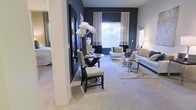 This single-floor space showcases a thoughtfully designed open-concept layout that seamlessly connects the dining and living areas. A striking dark accent wall anchors the dining space, creating a bold contrast against the lighter surrounding walls and plush neutral carpeting that runs throughout. The dining area features a sleek round glass-top table paired with contemporary dark-framed chairs with upholstered seats, accented by a tall decorative floral arrangement that adds an elegant focal point. Natural light filters in through a large window dressed with layered curtains in white and charcoal, enhancing the modern, curated atmosphere. The adjacent living area is furnished with a neutral-toned sofa and coordinating accent chairs arranged around a geometric area rug, complemented by slim metallic side tables. A secondary window on the far wall brings in additional natural light. Structural columns subtly define the transition between spaces while maintaining the open feel. The overall ambiance is sophisticated and contemporary, blending dark and light tones to create a visually dynamic yet inviting environment.