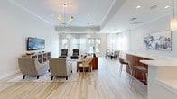 This well-appointed single-floor community space is designed with a modern aesthetic that balances comfort and functionality. Light hardwood-style flooring flows continuously throughout, creating a cohesive and airy atmosphere across all areas.

The open-plan layout connects a fully equipped kitchen, a casual dining nook, and a spacious lounge area. The kitchen stands out with rich walnut cabinetry, crisp white quartz countertops, a textured stone mosaic backsplash, stainless steel appliances, and pendant lighting that adds a warm, contemporary touch. A wraparound breakfast bar with bar stools provides an inviting casual seating option.

Adjacent to the kitchen, the main lounge area features a tray ceiling with a statement chandelier, recessed lighting, and large glass doors dressed in sheer white curtains that invite an abundance of natural light. The seating arrangement is thoughtfully laid out to encourage conversation, centered around a mounted flat-screen television. A separate dining area with a round table and seating for four sits beside floor-to-ceiling windows and direct access to an outdoor pool area.

Throughout the space, curated artwork and neutral-toned furnishings contribute to a polished yet welcoming environment. The overall design strikes a balance between upscale finishes and an approachable, communal character, making it an ideal gathering space.