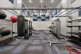 This fitness center is designed to accommodate a comprehensive range of workout modalities within a single, open-plan space. The room features notably high ceilings accented by a striking checkerboard pattern of blue and gray acoustic panels, which both enhance the aesthetic and manage sound levels throughout the space. A ceiling-mounted fan provides air circulation, complemented by recessed and pendant lighting that ensures the area is well-lit.

A prominent overhead monkey bar rig spans much of the ceiling, supporting functional training movements and serving as a defining architectural feature. Along one side of the room, full-length mirrors visually expand the space and assist users with form. The flooring consists of modular carpet tiles laid in a geometric pattern, providing a durable and cushioned surface suitable for various exercise types.

Equipment is thoughtfully distributed throughout. A dedicated strength training zone includes cable machines, adjustable benches, and a heavy bag with a medicine ball for combat-style training. A functional training station holds an organized rack stocked with kettlebells, medicine balls, and battle ropes. The cardio area offers ellipticals and rowing machines. Additional accessories such as stability balls and resistance equipment are neatly stored and accessible, making this fitness center a thoroughly outfitted training environment.