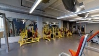 This fitness facility offers a thoughtfully organized workout environment spread across a single floor. The space features an industrial-style aesthetic, with exposed ductwork and piping running along the ceiling, complemented by bright fluorescent lighting that ensures excellent visibility throughout. The flooring consists of smooth, durable grey surfaces suited for heavy equipment and high-traffic use.

The strength training area is a standout feature, showcasing a comprehensive lineup of bold yellow-and-red resistance machines — including chest press, lat pulldown, leg press, and various other selectorized units — arranged in a spacious, accessible layout. Full-length mirrors line one wall, allowing users to monitor their form during workouts and adding a sense of depth to the room.

The open layout provides ample circulation space between equipment stations, making it easy for multiple users to train simultaneously without feeling crowded. Additional equipment is visible toward the back of the space, suggesting further workout options beyond the primary machine area. The overall ambiance is energetic and functional, with a color scheme that is visually striking and motivating. This facility is well-suited for strength training enthusiasts seeking a fully equipped, no-frills workout environment.