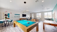 Located at 38 Westgate Dr. in Dover, NH, this well-appointed game room offers a generously sized, single-floor layout designed for recreation and social engagement. The space is anchored by a striking pool table with a vivid teal felt surface and a solid wood frame, positioned beneath a contemporary circular pendant light fixture that serves as a bold design centerpiece. Complementing the pool table are a foosball table, a full-size ping pong table, and a shuffleboard table, providing a diverse array of gaming options. High-top tables paired with teal upholstered bar chairs are arranged throughout the room, offering comfortable spots for socializing between games. A wall-mounted flat-screen TV adds an entertainment focal point to the space. The interior design features light wood-look flooring, crisp white ceilings with recessed lighting, and a bold teal accent wall that injects vibrant color and personality into the room. Multiple windows along the exterior walls invite abundant natural light and provide views to the outdoors. A pass-through window connects to an adjacent kitchen area, adding functional convenience. The overall ambiance is modern, lively, and well-suited for community gatherings and casual entertainment.
