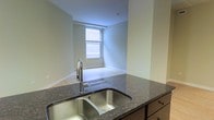 This single-floor space offers a clean, modern layout designed for comfortable everyday living. The kitchen is a standout feature, showcasing dark granite countertops with subtle natural stone patterning and a double-basin stainless steel sink with a sleek pull-down faucet. Rich, dark wood cabinetry complements the countertops, adding warmth and ample storage to the space. The kitchen opens directly to an adjacent living area, creating an easy flow between spaces. The living area is bathed in natural light courtesy of a large window fitted with horizontal blinds, allowing for adjustable brightness throughout the day. Light-toned flooring in the living area provides a pleasing contrast to the darker kitchen finishes. The walls throughout are finished in a soft, neutral green-grey tone, contributing to a calm and understated ambiance. White baseboards offer a crisp finishing detail throughout. A smoke detector is visible on the ceiling, reflecting thoughtful safety considerations. Overall, the space presents a well-maintained, move-in-ready interior with a cohesive design palette that balances contemporary finishes with functional layout choices.