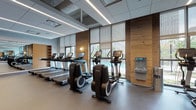This fitness center is a spacious, single-floor facility designed to support a comprehensive range of workout routines. The space is thoughtfully organized into distinct zones, beginning with a well-stocked cardio area lined with treadmills and elliptical machines, each equipped with individual digital screens for interactive training. Adjacent to this section, a dedicated cycling area features stationary bikes, including Peloton-style units, positioned along floor-to-ceiling windows that frame outdoor views and flood the space with natural light. A stair climber rounds out the cardio offerings. The strength and functional training zone provides a cable machine system, free weights, and a dumbbell rack, complemented by full-length mirrors that enhance the sense of depth throughout the room. A built-in water bottle refill station is conveniently located near the cardio section. The interior design balances modern and natural elements, with warm wood accent panels contrasting against a neutral palette of grey flooring and a suspended tile ceiling with sleek linear lighting. The open floor plan allows for ample movement space, while roller shades on the large windows offer adjustable light control. The overall ambiance is clean, bright, and professionally appointed.
