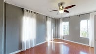 This single-floor space presents a clean, well-lit interior designed for both comfort and functionality. The main living area features warm-toned hardwood flooring that runs throughout, reflecting natural light and adding a sense of warmth to the room. The walls are finished in a soft gray tone, providing a neutral and modern backdrop that complements the overall aesthetic.

Large windows dressed with sheer white curtains line multiple walls, allowing an abundance of natural light to pour into the space while maintaining a light, airy feel. The curtains are hung on rod-and-ring systems, adding a subtle decorative touch. One window offers an outward view of surrounding greenery, connecting the interior to the outdoors.

A five-blade ceiling fan with an integrated light fixture is centrally mounted, ensuring effective air circulation and supplemental lighting throughout the room. White baseboards provide a crisp contrast against the gray walls and hardwood floors, contributing to a polished, cohesive finish. Crown molding detail at the ceiling further enhances the refined character of the space. Overall, the room conveys a bright, open, and move-in-ready atmosphere.