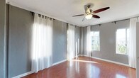 This single-floor space presents a clean, well-lit interior designed for both comfort and functionality. The main living area features warm-toned hardwood flooring that runs throughout, reflecting natural light and adding a sense of warmth to the room. The walls are finished in a soft gray tone, providing a neutral and modern backdrop that complements the overall aesthetic.

Large windows dressed with sheer white curtains line multiple walls, allowing an abundance of natural light to pour into the space while maintaining a light, airy feel. The curtains are hung on rod-and-ring systems, adding a subtle decorative touch. One window offers an outward view of surrounding greenery, connecting the interior to the outdoors.

A five-blade ceiling fan with an integrated light fixture is centrally mounted, ensuring effective air circulation and supplemental lighting throughout the room. White baseboards provide a crisp contrast against the gray walls and hardwood floors, contributing to a polished, cohesive finish. Crown molding detail at the ceiling further enhances the refined character of the space. Overall, the room conveys a bright, open, and move-in-ready atmosphere.