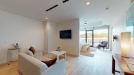This thoughtfully designed single-floor unit combines contemporary aesthetics with practical comfort across a seamlessly flowing open layout. Light hardwood flooring runs throughout the main living areas, tying together the living, dining, and sleeping zones with visual cohesion.

The living area features a plush sectional sofa, a wall-mounted flat-screen TV, and recessed lighting, creating an inviting space for relaxation. It flows directly into a compact but fully equipped kitchen outfitted with wood-toned cabinetry, stainless steel appliances including a French door refrigerator and gas range, a subway tile backsplash, and a glass-top dining table with upholstered chairs.

The sleeping area is defined by a tray ceiling and anchored by a generously sized bed with an upholstered headboard, flanked by navy nightstands and warm table lamps. A ceiling fan adds comfort, and floor-to-ceiling glass doors open directly onto a private balcony with outdoor seating and tree-lined views, filling the room with natural light.

The bathroom is finished in a clean, modern style with large-format tile flooring, a wood-grain vanity with a white quartz countertop, an undermount sink, and a tub-shower combination. The space as a whole presents a refined, neutral palette with carefully selected furnishings that balance style and function.