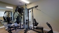 This single-floor fitness space is thoughtfully designed to serve as a fully functional home gym. The room is anchored by a large multi-station cable machine, which offers a wide variety of strength-training exercises including lat pulldowns, cable rows, and other resistance-based movements. An adjustable workout bench is positioned nearby, adding further versatility for weight training routines. A cardio machine, visible in the mirror reflection, rounds out the equipment selection and provides options for cardiovascular exercise alongside strength training. The walls are fitted with full-length mirrors, which not only create a sense of expanded space but also allow users to monitor their form during workouts. The room features neutral-toned walls and carpeted flooring, contributing to a clean and functional aesthetic. A window allows for natural light to filter into the space, enhancing the overall environment. The ceiling features a fluorescent light fixture that ensures the room remains well-lit at all times. The layout is efficient, making good use of the available floor area while keeping the space organized and accessible for a comprehensive fitness routine.