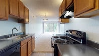 This single-floor unit offers a practical and well-organized layout designed for comfortable everyday living. The kitchen is equipped with warm honey-toned wood cabinetry, dark countertops, and black appliances including a coil-burner electric range and a full-size refrigerator. Wood-look vinyl flooring runs through the kitchen, and a range hood provides ventilation above the stove. The kitchen flows directly into the living area, which is finished with plush neutral carpeting and features a striking gray accent wall. A sliding glass door leads to a private balcony, bringing in generous natural light and offering views of lush greenery beyond. Baseboard heating lines the perimeter of the living space, ensuring warmth throughout. A pendant light fixture adds a subtle decorative touch to the open kitchen-living transition. The bathroom is thoughtfully appointed with a white vanity featuring dark countertops and a mirrored medicine cabinet, wood-look tile flooring, and a full tub-shower combination. Textured ceilings are present throughout the unit, adding a classic touch. Overall, the space presents a clean, functional, and move-in ready environment with a cohesive neutral palette.