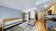 This single-floor studio unit offers a thoughtfully organized open-plan layout that efficiently combines living, sleeping, and kitchen functions within one cohesive space. The sleeping area features a twin bed with a trundle, providing flexible sleeping arrangements, and is positioned on durable carpet tiles that define the zone. A built-in closet with open shelving and a gray-toned door offers generous wardrobe and storage solutions. An accent armchair provides a comfortable seating nook, complementing the relaxed ambiance of the room. The flooring transitions from carpet in the sleeping area to warm hardwood-style planks that extend toward the kitchenette and entryway, adding visual depth and continuity. Natural light streams in through a window, casting long sunlit streaks across the floor and enhancing the brightness of the space. The kitchenette area is equipped with a full-size refrigerator and countertop space with cabinetry, offering practical day-to-day functionality. Recessed ceiling lighting throughout ensures consistent illumination. The light gray and neutral wall tones, combined with the warm wood accents of the furniture and cabinetry, create a clean, modern, and welcoming interior atmosphere well-suited for comfortable everyday living.