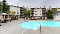 This residential community offers an impressive array of shared outdoor amenities designed for comfort and recreation. At the heart of the property is a generously sized, kidney-shaped swimming pool with crystal-clear blue water, surrounded by a wide, textured concrete sundeck. The deck is well-furnished with a selection of lounge chairs and seating areas, providing ample space for relaxing or socializing in the open air.

Adjacent to the pool area sits a dedicated clubhouse building with a welcoming exterior and large glass-paned doors. An outdoor patio extends from the clubhouse entrance, complete with umbrella-shaded dining tables and chairs — ideal for casual gatherings. Mature evergreen trees and carefully maintained landscaping frame the entire outdoor space, adding a sense of privacy and a natural, Pacific Northwest ambiance.

A standout feature of the community is the covered outdoor grilling station, tucked beneath a wood pergola. The built-in stainless steel grill is set against a striking backsplash of multi-toned slate tiles arranged in a diamond pattern, flanked by stone-clad pillars — a thoughtful and stylish detail that elevates the outdoor cooking experience.

The surrounding multi-story residential buildings feature private balconies and neutral exterior siding, contributing to the cohesive and well-maintained character of the community.