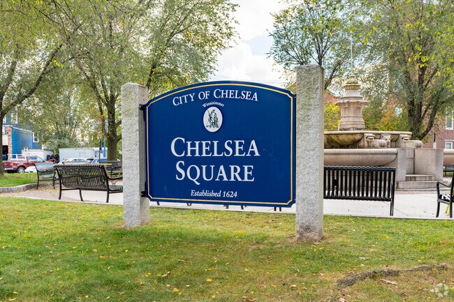 Chelsea Square sign stands in the middle of a park with a water fountain, benches, and shops.