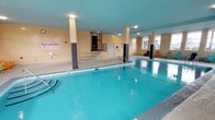 This single-level indoor aquatic facility is defined by its generously sized swimming pool, which serves as the centerpiece of the space. The pool features clearly marked depth indicators and is equipped with stainless steel entry railings, ensuring safe and convenient access for swimmers. A wheelchair lift platform is also present, underscoring the facility's commitment to accessibility. Surrounding the pool, the deck area features slip-resistant flooring, providing a safe walking surface throughout.

The walls are constructed of painted concrete masonry block in a warm beige tone, complemented by a distinctive teal-painted ceiling that adds a vibrant, aquatic atmosphere to the interior. Multiple rows of large windows line the perimeter walls, allowing substantial natural light to fill the space while offering views of the surrounding outdoor greenery and landscape.

Designated relaxation areas are furnished with colorful Adirondack chairs and small side tables, arranged along the windowed walls to create comfortable poolside lounging spots. Practical amenities include posted facility notices, a wall-mounted clock, fire alarm systems, and ventilation units. Staircase access with stainless steel handrails connects different sections of the facility, further enhancing circulation and usability throughout this well-appointed aquatic space.
