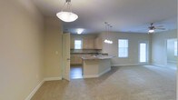 This single-floor space offers a clean, open-concept layout that connects the main living area to the kitchen with a natural, uninterrupted flow. The living area is finished with wall-to-wall carpet in a neutral tone, creating a comfortable and cohesive atmosphere throughout.

The kitchen is a central highlight, featuring light wood cabinetry, dark granite countertops, and a peninsula island that provides additional counter space and serves as a natural dividing point between the kitchen and living areas. A stainless steel sink is positioned along the kitchen counter, and the space is illuminated by a stylish multi-bulb pendant light fixture that hangs above the island, complementing a matching pendant in the main living area.

Natural light enters through multiple windows fitted with blinds, brightening the neutral beige and gray wall tones and giving the interior a fresh, airy feel. The kitchen includes tile flooring that transitions into the carpeted living space, clearly delineating the two functional zones. A doorway off the kitchen leads to an additional room, offering further utility and flexibility within the layout. Overall, the space presents a practical and well-lit environment with a consistent, understated aesthetic throughout.