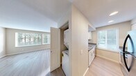 Located at 3030 NE 143rd St. in Seattle, WA, this well-appointed one-bedroom, one-bathroom unit offers a practical and comfortable single-floor layout. The open-concept kitchen and living area serves as the heart of the home, featuring light wood-style flooring, neutral warm-toned walls, and an abundance of natural light streaming through large windows dressed with horizontal blinds. The kitchen is equipped with white shaker-style cabinetry, stainless steel appliances including a range and refrigerator, and a peninsula with a double sink and gray countertops — ideal for both cooking and casual dining. A dedicated closet off the main living area houses a stacked washer and dryer, offering the convenience of in-unit laundry. The bedroom is a generously sized, carpet-floored retreat with a large window overlooking lush greenery, providing a tranquil and private atmosphere. A dedicated closet offers ample storage. The full bathroom features a clean, white tub/shower combo with chrome fixtures and a simple, functional design. Throughout the unit, clean lines, consistent neutral tones, and plentiful natural light create a bright and cohesive living environment.