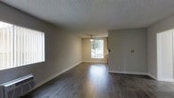 This single-floor residence offers a clean, functional layout with a welcoming and well-lit interior. The main living area is a standout feature, showcasing rich, dark hardwood-style flooring that runs throughout and reflects the natural light that pours in through a large sliding glass door. The door provides views of the outdoors and serves as both a practical entry point and a bright focal point for the room. A ceiling fan is centrally positioned overhead, offering air circulation and a subtle decorative touch. The walls are finished in a neutral, warm beige tone that complements the flooring and creates a cohesive, understated aesthetic. A textured popcorn ceiling adds a classic character to the space. White baseboards provide a crisp contrast against the walls and floors, contributing to a tidy, polished look. Additional windows along the side walls further enhance the natural light, keeping the space feeling open and airy. The overall ambiance is calm and practical, making it a versatile space well-suited for a variety of furnishing styles and uses.