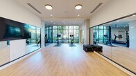 Located at 3010 Edgeview Ln, this single-floor fitness center spans 2,230 square feet of thoughtfully designed workout space. The facility is divided into distinct functional zones, each tailored to a specific training style. The main strength and cardio area features textured loop carpet flooring, recessed lighting, and ceiling fans, creating a comfortable environment for extended workouts. This zone is equipped with a multi-station cable machine, weight benches, treadmills, stair climbers, and ellipticals, complemented by a wall-mounted TV and built-in bench seating with storage. Large floor-to-ceiling windows flood the space with natural light and provide views of the surrounding greenery. A dedicated group fitness and cycling studio boasts light hardwood floors, wall-to-wall mirrors, stationary bikes with integrated screens, and expansive black-framed glass walls that open the space visually to the outdoors. A separate functional training room offers hardwood flooring, mirrored walls, a wall-mounted rig with a heavy punching bag, medicine balls, resistance tools, and step platforms—ideal for dynamic and combat-style training. A vibrant accent wall featuring a bold hexagonal mosaic pattern adds a distinctive design element to the main gym area, giving the overall facility a polished, contemporary aesthetic.