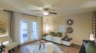 This single-floor space offers a thoughtfully designed living area that balances comfort and style. The living room serves as the heart of the home, featuring rich hardwood flooring that adds warmth and character throughout. A ceiling fan with an integrated light fixture is centrally positioned, providing both air circulation and ambient lighting. Natural light pours generously into the room through elegant French doors adorned with flowing white curtains, offering a seamless connection to the outdoors. The living area is anchored by a neutral-toned sofa dressed with colorful accent pillows, complemented by decorative wall art and sunburst wall decor that adds a touch of organic texture and visual interest. A tall woven floor lamp provides additional warm lighting, enhancing the cozy atmosphere. The space is finished with a cowhide area rug layered over the hardwood floors, adding a stylish, eclectic touch. The overall ambiance is light, airy, and welcoming, with a warm neutral color palette that creates an inviting and relaxed environment throughout.