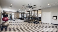 Located in Metairie, Louisiana, this fitness center at Lumiere offers a thoughtfully designed, single-floor workout space that balances functionality with modern aesthetics. The standout design feature is the striking chevron-patterned flooring in neutral tones of gray, white, and taupe, which lends the space a sophisticated, contemporary feel throughout.

The center is generously equipped with a suite of True-brand cardio machines, including multiple treadmills, ellipticals, and stationary bikes, arranged to maximize floor space and ease of movement. A multi-station strength machine and a dedicated free-weight area round out the equipment offerings, with organized racks holding medicine balls, stability balls, and kettlebells in a variety of sizes. Colorful abstract artwork adorns the walls, adding visual interest to the workout environment.

Large-blade ceiling fans provide consistent air circulation, complementing the recessed lighting overhead. Arched windows along one wall invite natural light and are fitted with blinds for adjustable comfort. Floor-to-ceiling glass panel walls and doors connect the fitness center to an adjacent common area, creating a sense of openness. A posted fitness center rules board, a water dispenser, and a waste receptacle near the entrance ensure a clean, well-managed facility. The overall ambiance is polished and welcoming, designed to support a wide range of fitness routines.