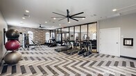 Located in Metairie, Louisiana, this fitness center at Lumiere offers a thoughtfully designed, single-floor workout space that balances functionality with modern aesthetics. The standout design feature is the striking chevron-patterned flooring in neutral tones of gray, white, and taupe, which lends the space a sophisticated, contemporary feel throughout.

The center is generously equipped with a suite of True-brand cardio machines, including multiple treadmills, ellipticals, and stationary bikes, arranged to maximize floor space and ease of movement. A multi-station strength machine and a dedicated free-weight area round out the equipment offerings, with organized racks holding medicine balls, stability balls, and kettlebells in a variety of sizes. Colorful abstract artwork adorns the walls, adding visual interest to the workout environment.

Large-blade ceiling fans provide consistent air circulation, complementing the recessed lighting overhead. Arched windows along one wall invite natural light and are fitted with blinds for adjustable comfort. Floor-to-ceiling glass panel walls and doors connect the fitness center to an adjacent common area, creating a sense of openness. A posted fitness center rules board, a water dispenser, and a waste receptacle near the entrance ensure a clean, well-managed facility. The overall ambiance is polished and welcoming, designed to support a wide range of fitness routines.