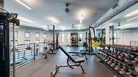 Located at 5700 Crossings Blvd in Nashville, Tennessee, this fitness center offers a generously sized, single-floor exercise facility designed to support a full range of workout routines. The space features a modern industrial aesthetic, with exposed ductwork running along the ceiling, sleek linear lighting, and ceiling fans that maintain comfortable airflow throughout. Durable rubber flooring covers the workout areas, providing both safety and functionality.

The strength training zone is well-appointed with a power rack loaded with weight plates, a comprehensive dumbbell rack ranging across multiple weights, adjustable workout benches, and a cable machine. A large wall mirror enhances the space, allowing for form checks during exercises. A dedicated equipment storage rack holds medicine balls, resistance tools, and accessories for varied training.

The cardio section features multiple treadmills, an elliptical machine, and a stationary spin bike, all positioned near a row of windows that bring in natural light and outdoor views. A separate entry area includes a water station with a bottle-fill dispenser, a storage cubby unit, and an air purifier, adding convenience and practicality. A metal railing staircase connects to adjacent areas of the building. The overall ambiance is clean, modern, and motivating, making it a fully functional fitness destination.