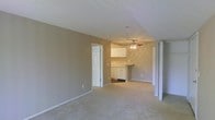 This single-floor residence offers a straightforward and functional layout designed for comfortable everyday living. The main living area is generously proportioned, featuring soft, neutral-toned walls and wall-to-wall carpeting that lends a warm, understated feel to the space. The open layout creates a seamless visual connection between the living area and the kitchen beyond, making the space feel airy and cohesive.

The kitchen is positioned at the far end of the main living area, outfitted with white cabinetry that provides ample storage. A ceiling fan is mounted above, offering both practical ventilation and a subtle decorative touch. The kitchen's lighter tones contrast pleasantly with the warm beige walls shared throughout the residence.

Doorways visible from the main living space suggest access to additional rooms, including what appears to be a closet with bifold doors and a passage to other areas of the home. The consistent use of white trim and baseboards throughout the space ties the design together with a clean, classic finish.

Overall, the space presents a calm, neutral palette and an efficient single-level floor plan that offers a practical and livable environment, well-suited for those seeking a simple, move-in-ready home.