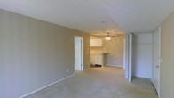 This single-floor residence offers a straightforward and functional layout designed for comfortable everyday living. The main living area is generously proportioned, featuring soft, neutral-toned walls and wall-to-wall carpeting that lends a warm, understated feel to the space. The open layout creates a seamless visual connection between the living area and the kitchen beyond, making the space feel airy and cohesive.

The kitchen is positioned at the far end of the main living area, outfitted with white cabinetry that provides ample storage. A ceiling fan is mounted above, offering both practical ventilation and a subtle decorative touch. The kitchen's lighter tones contrast pleasantly with the warm beige walls shared throughout the residence.

Doorways visible from the main living space suggest access to additional rooms, including what appears to be a closet with bifold doors and a passage to other areas of the home. The consistent use of white trim and baseboards throughout the space ties the design together with a clean, classic finish.

Overall, the space presents a calm, neutral palette and an efficient single-level floor plan that offers a practical and livable environment, well-suited for those seeking a simple, move-in-ready home.