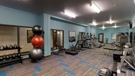 This fitness center offers a comprehensive workout environment designed to accommodate a variety of exercise routines. The space is finished with cool blue-toned walls that create an energizing atmosphere, complemented by patterned carpet flooring with blue accent inlays that adds a modern, dynamic feel underfoot. Overhead, flush-mounted fluorescent lighting ensures the entire area is brightly and evenly illuminated. The cardio section is well-appointed with multiple treadmills, elliptical machines, and a stationary air bike, all arranged in a row to maximize floor space and ease of use. A dedicated functional training area features an adjustable weight bench and a rack of exercise balls in multiple sizes, providing options for strength, balance, and flexibility training. Framed artwork displayed along the walls adds a welcoming touch to the otherwise utilitarian setting. The layout is open and spacious, allowing for comfortable movement between equipment. A doorway visible at the far end of the room suggests additional amenities or access points nearby, contributing to the overall convenience and accessibility of the facility. The space is thoughtfully organized to support a full range of fitness activities in a clean, motivating environment.